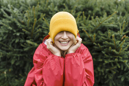 Lächelnde Frau in Regenmantel und Strickmütze vor Pflanzen im Park - VBUF00489