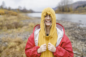 Fröhliche Frau, die ihr Kapuzenshirt vor einem Fluss anzieht - VBUF00494