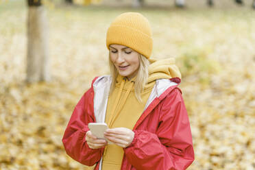 Lächelnde Frau in rotem Regenmantel und mit Smartphone im Park - VBUF00498