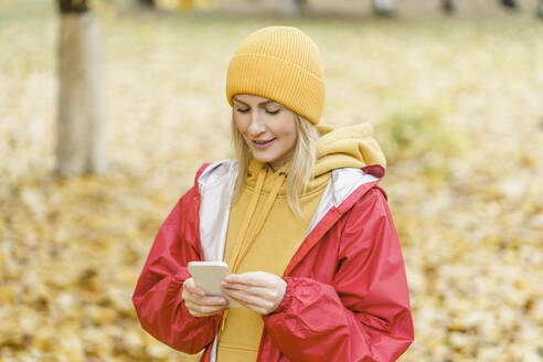 Lächelnde Frau in rotem Regenmantel und mit Smartphone im Park - VBUF00498