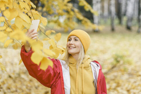 Smiling woman wearing yellow knit hat and taking selfie through smart phone in park - VBUF00499