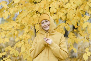 Happy woman wearing hooded shirt and knit hat in front yellow leaves at park - VBUF00500