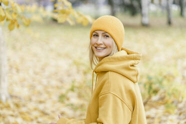 Happy woman wearing yellow hooded shirt and knit hat in park - VBUF00501