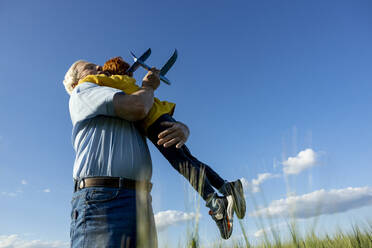 Elderly man with toy airplane embracing grandson in field under sky - MBLF00096