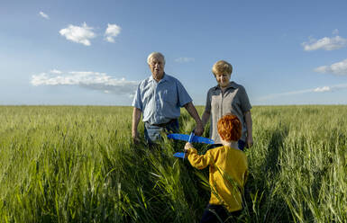 Grandson holding toy airplane spending leisure time with grandparents in field - MBLF00101