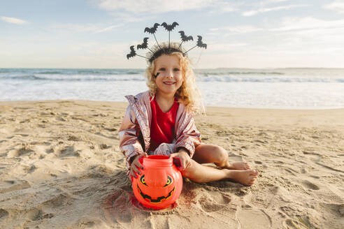 Lächelndes Mädchen sitzt mit Halloween-Kürbis Spielzeug auf Sand am Strand - SIF01065