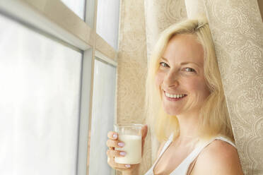 Lächelnde Frau mit einem Glas Milch am Fenster zu Hause - VGF00426