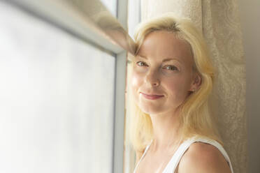 Smiling blond woman leaning on window at home - VGF00427