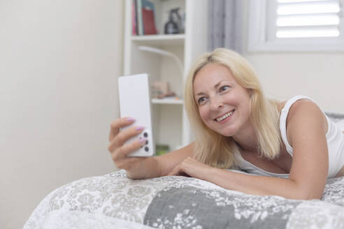 Glückliche Frau führt einen Videoanruf über ihr Smartphone durch, während sie zu Hause auf dem Bett liegt - VGF00428