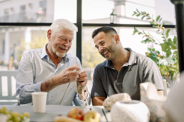 Happy male nurse sitting with senior man crocheting at nursing home - MASF40184