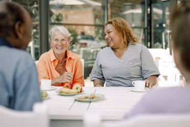 Cheerful young female nurse sitting with retired senior man and women at dining table - MASF40193