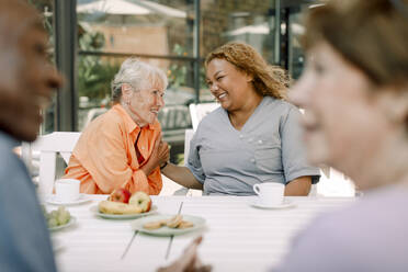 Cheerful young female caregiver sitting with retired senior man and women at dining table - MASF40194