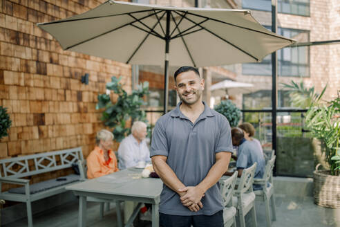 Portrait of smiling male nurse standing with hands clasped at nursing home - MASF40204