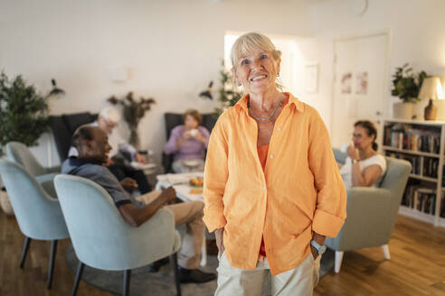 Portrait of smiling retired senior woman standing with hands in pockets at nursing home - MASF40208