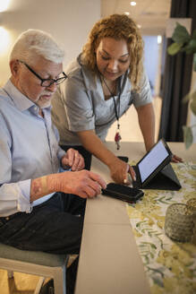 Young female caregiver assisting senior man in using smart phone at retirement home - MASF40214