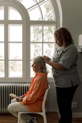 Side view of young female caregiver braiding senior woman's hair at nursing home - MASF40218
