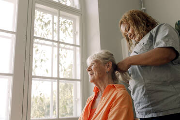 Young female caregiver braiding smiling senior woman's hair at nursing home - MASF40220