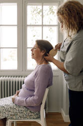 Side view of young female nurse brushing senior woman's hair at retirement home - MASF40222