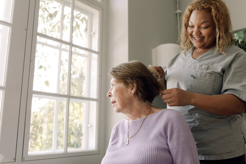 Low angle view of young female nurse brushing senior woman's hair at retirement home - MASF40223
