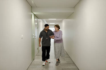 Full length of smiling senior woman walking arm in arm with male nurse at retirement home corridor - MASF40227