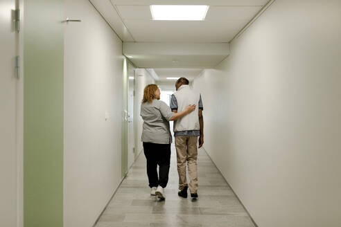 Full length rear view of female nurse walking with arm around senior man in corridor at retirement home - MASF40229