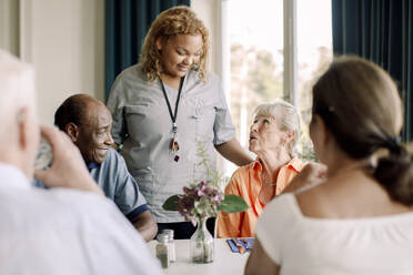 Smiling young female caregiver talking to senior women and men at dining table in nursing home - MASF40232