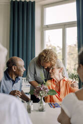 Young female caregiver embracing senior woman sitting with friends at dining table in nursing home - MASF40233