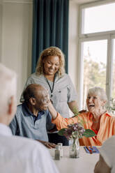 Smiling young female caregiver standing by happy senior women and men at dining table in nursing home - MASF40234