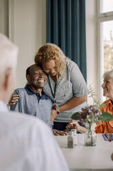 Smiling young female caregiver embracing happy senior men sitting with friends at dining table in nursing home - MASF40235