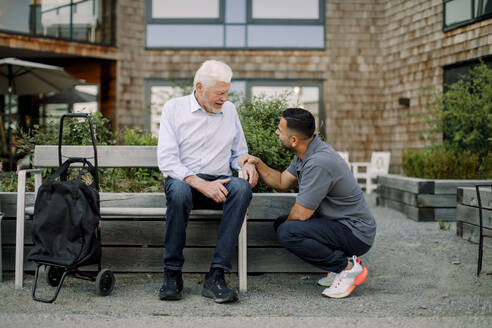 Full length of male nurse squatting while talking to retired senior man sitting on bench at back yard - MASF40236