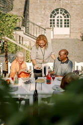 Smiling female caregiver serving food to senior man and woman sitting at dining table in back yard - MASF40243