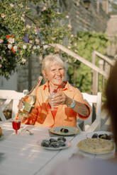 Senior woman holding alcohol bottle while sitting at dining table in back yard - MASF40246