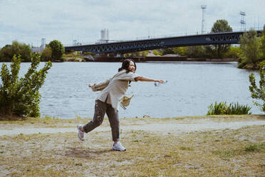 Full length of young woman throwing water from bottle near river - MASF40259