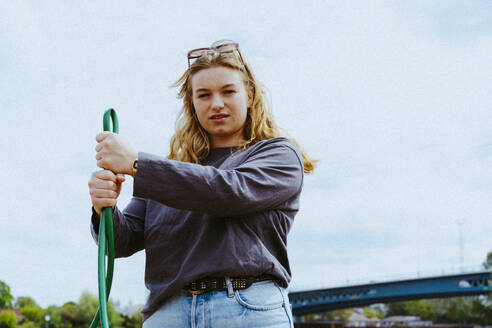 Low angle portrait of confident young blond woman holding garden hose against sky - MASF40263