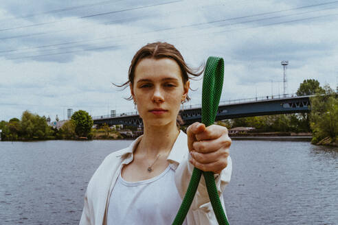 Porträt einer selbstbewussten jungen nicht-binären Person, die einen Gartenschlauch in der Nähe eines Flusses hält - MASF40274