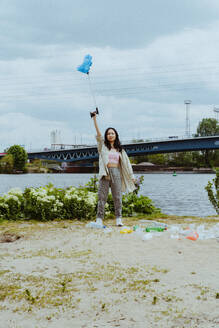 Full length portrait of confident young woman picking garbage near river - MASF40284