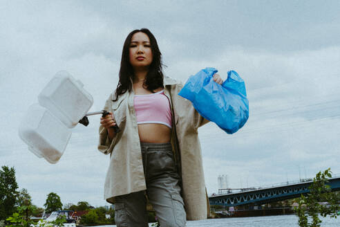 Low angle portrait of confident woman picking garbage near river - MASF40286