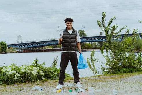 Portrait of smiling young man collecting garbage near river against sky - MASF40288