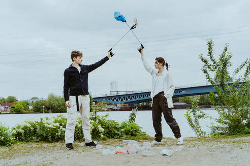 Young friends collecting plastic garbage near river against sky - MASF40289