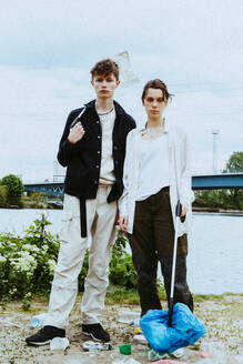 Full length portrait of young friends collecting plastic garbage near river against sky - MASF40291