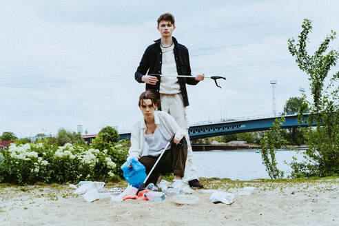 Full length portrait of confident young friends collecting plastic garbage near river against sky - MASF40292