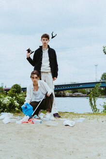 Full length portrait of confident young friends collecting plastic waste near river against sky - MASF40293