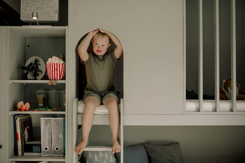 Full length of boy with down syndrome sitting on bunkbed ladder at home - MASF40308
