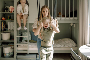 Mother helping son with down syndrome swinging on gymnastics rings in bedroom - MASF40309