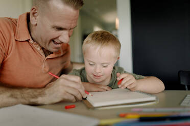 Father assisting son with down syndrome in writing on book at dining table - MASF40320