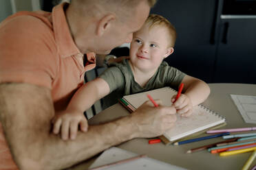 Boy with down syndrome looking at father while drawing on book - MASF40321