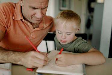 Father and son with down syndrome drawing together in book at dining table - MASF40322