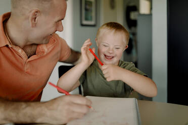 Happy father and son with down syndrome drawing in book at dining table - MASF40323