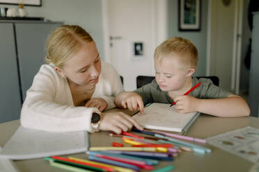 Girl assisting brother with down syndrome in drawing on book at home - MASF40324