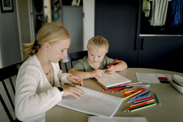 Girl doing homework by brother with down syndrome drawing in book at home - MASF40325